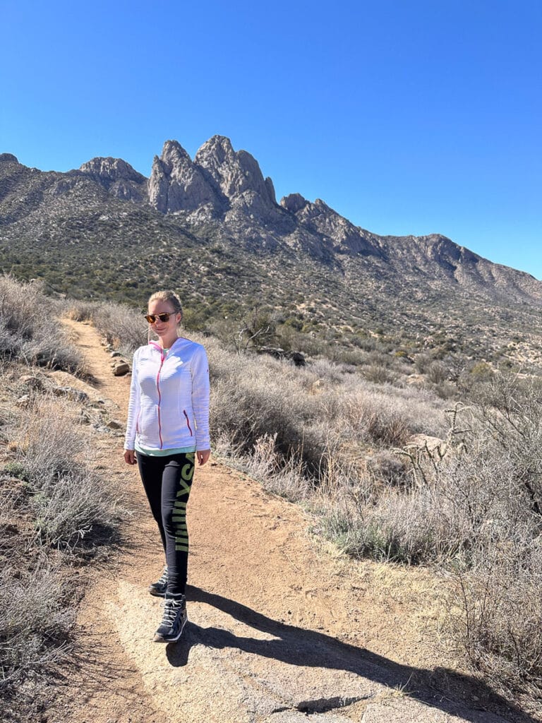 The author stands on a dirt trail with the mountain range behind her. She is dressed in sporty attire, ready for a hike, with a confident stance and sunglasses on.