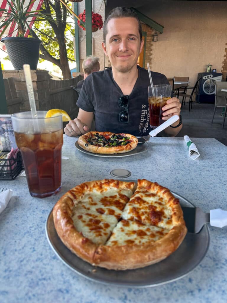 A cozy patio meal at an Alamogordo restaurant. The foreground displays a personal pizza topped with melted cheese, with iced tea in a tall glass. The background shows another pizza, highlighting the restaurant's comforting ambiance.