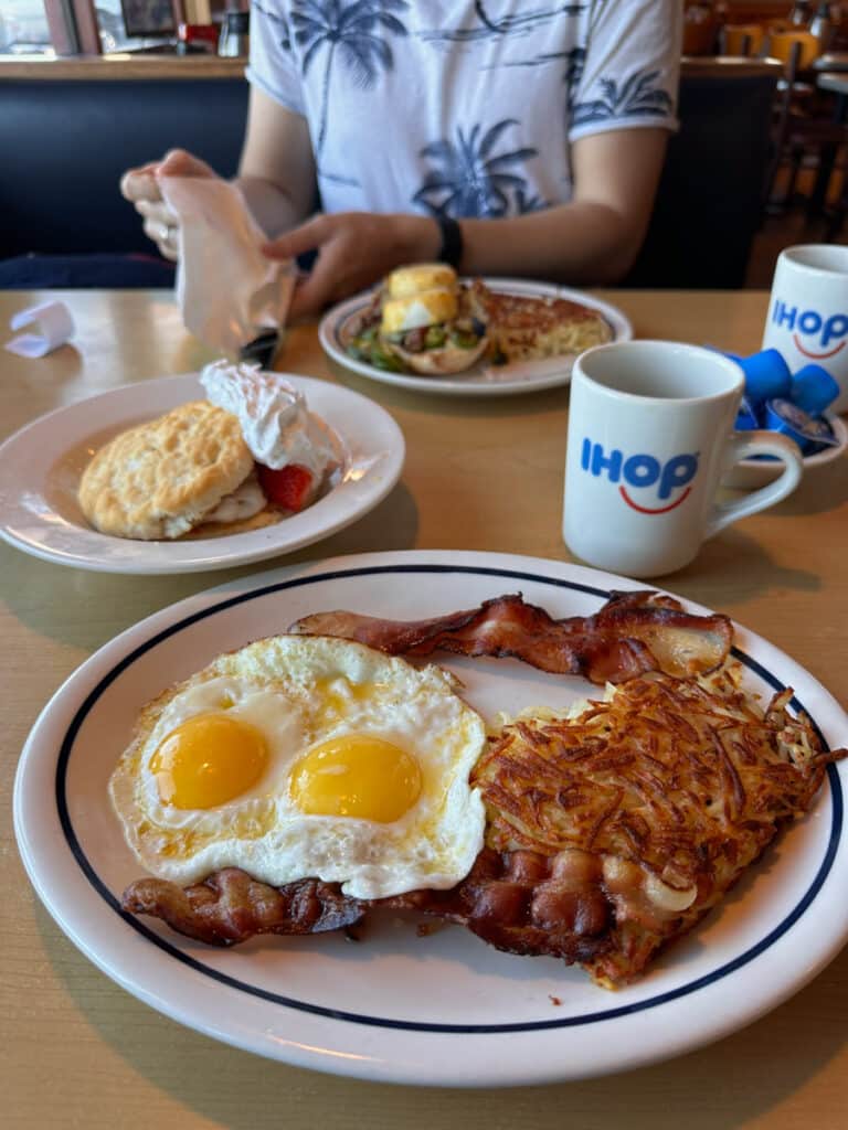A delicious breakfast plate at IHOP, featuring sunny-side-up eggs, crispy bacon, golden hash browns, and two sausage links. Nearby, a cup of coffee with the IHOP logo, and a second breakfast plate in the background, completing the classic American breakfast scene at one of the best Alamogordo restaurants.